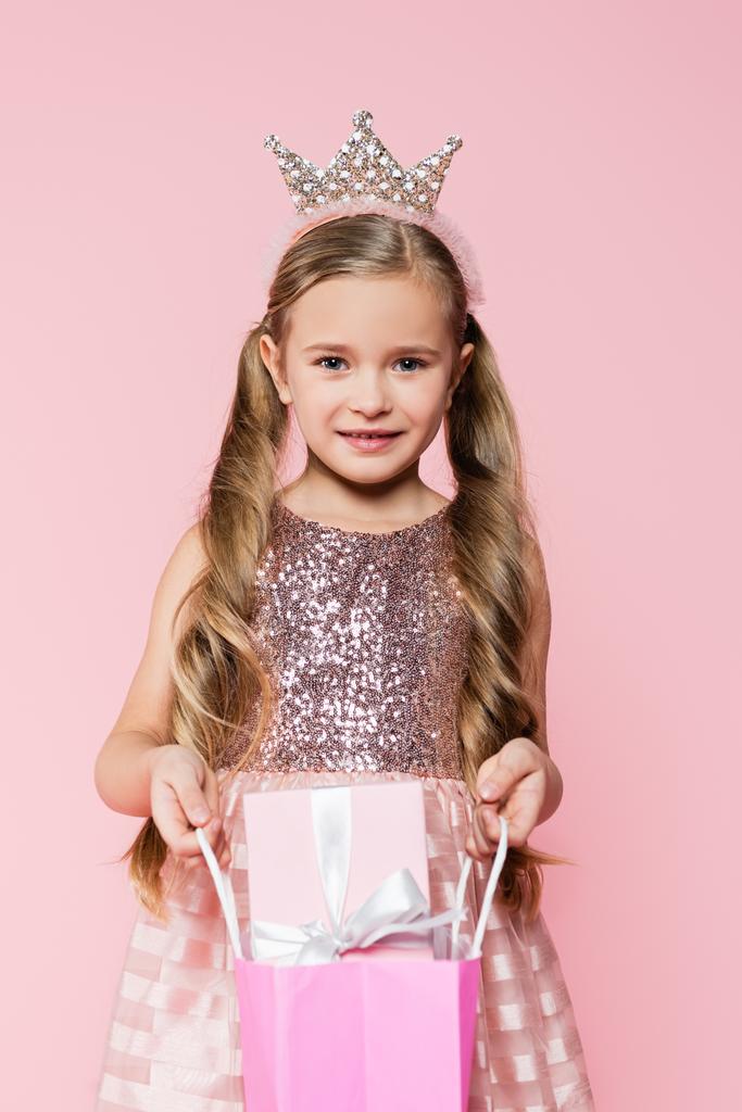 boldog kislány ruha és korona gazdaság bevásárló táska ajándék elszigetelt rózsaszín  - Fotó, kép