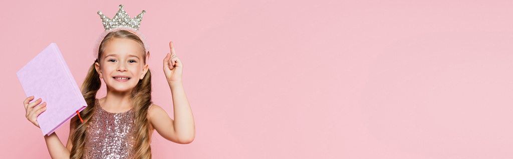 χαμογελαστό κοριτσάκι στο βιβλίο κρατώντας στέμμα και δείχνοντας με το δάχτυλο απομονώνονται σε ροζ, banner - Φωτογραφία, εικόνα