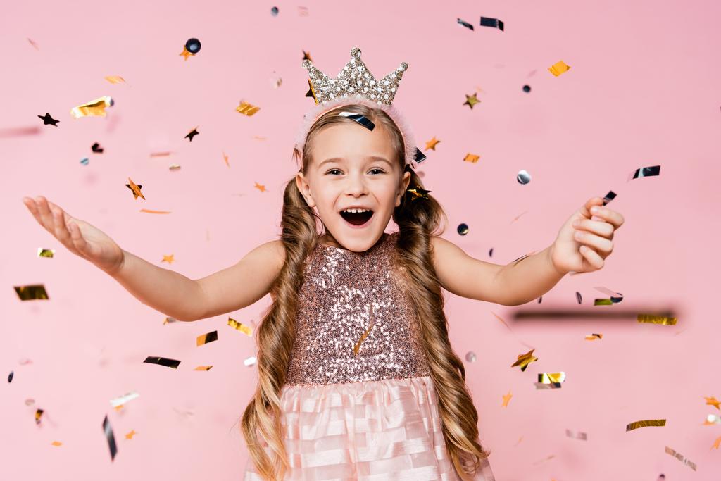zdumiona mała dziewczynka w koronie z wyciągniętymi rękami w pobliżu spadającego konfetti na różowy  - Zdjęcie, obraz