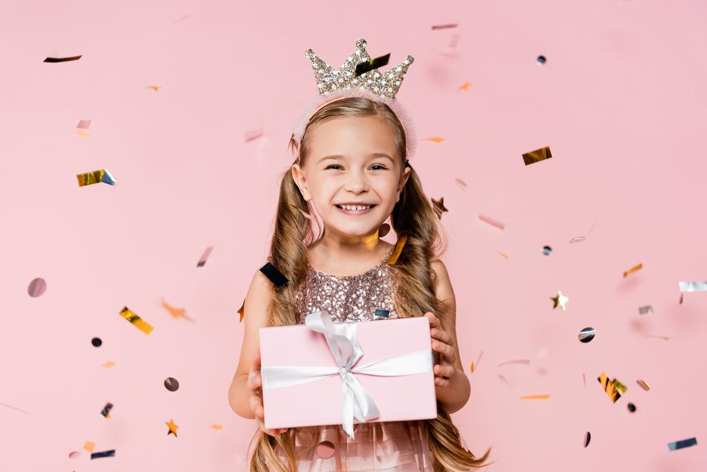 happy little girl in crown holding present near falling confetti on pink  - Photo, Image