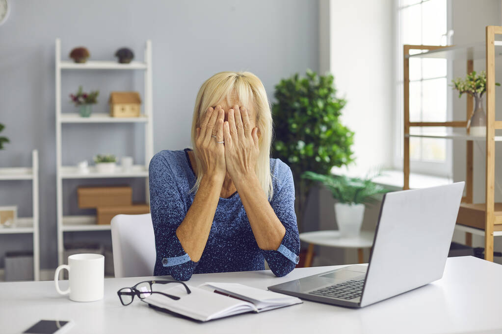 Mujer mayor molesto sentado en el escritorio con el ordenador portátil y la cara cubierta incapaz de hacer frente a los problemas - Foto, Imagen