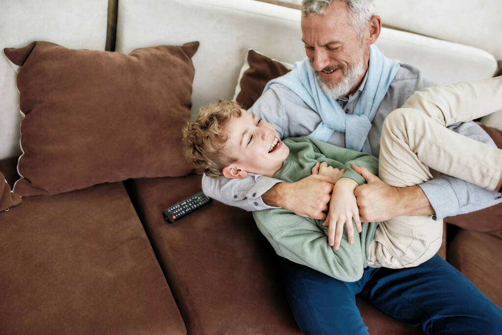 Nejlepší přátelé. Portrét veselého dědečka a vzrušeného vnuka objímající, hrající a bavící se při relaxaci na gauči doma - Fotografie, Obrázek