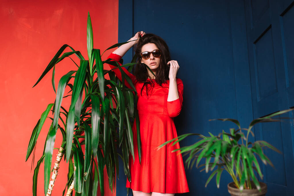 Красива дівчина моди з довгим волоссям, в сонцезахисних окулярах і червона елегантна сукня позує на синій червоній стіні між зеленими деревами для камери. Внутрішній м'який фокус портрет стильної пристрасної брюнетки красуня
 - Фото, зображення