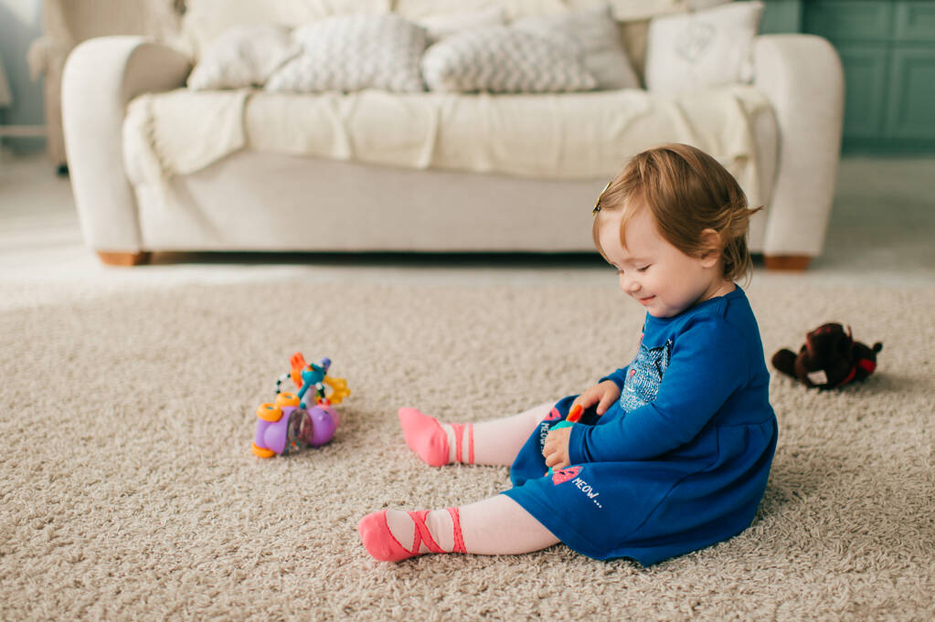 Μικρό χαριτωμένο καυκάσιο κορίτσι σε ελκυστικό φόρεμα κάθεται στο πάτωμα και χαλί, παίζει με διάφορα παιχνίδια και χαμόγελα της - Φωτογραφία, εικόνα