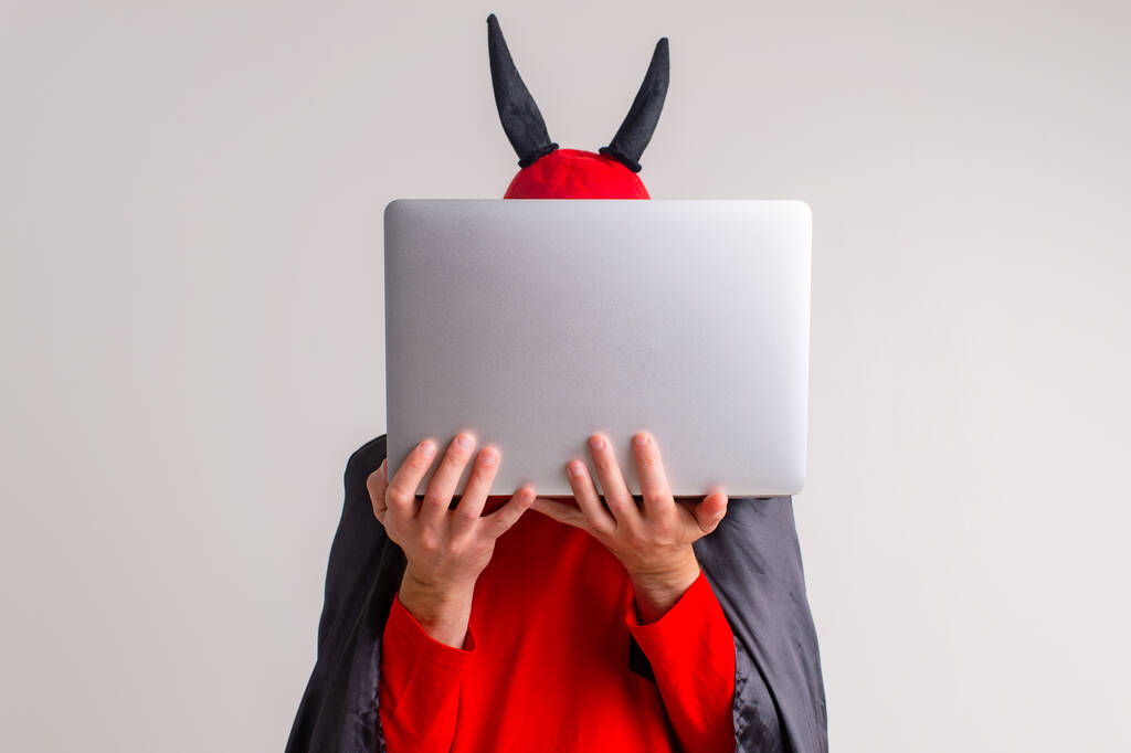 Stylowy biały mężczyzna w kapeluszu diabła z rogami i peleryną wampira z laptopem izolowane na białym backhround. - Zdjęcie, obraz