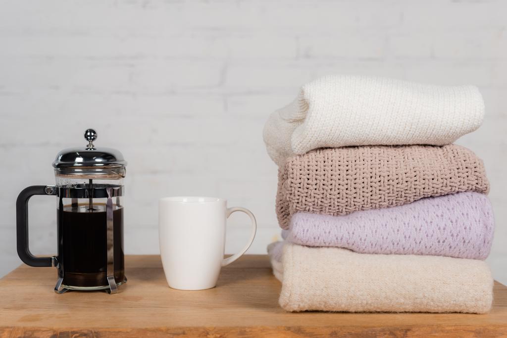 Чашка и чайник возле вязаных свитеров на столе на фоне кирпичной стены - Фото, изображение