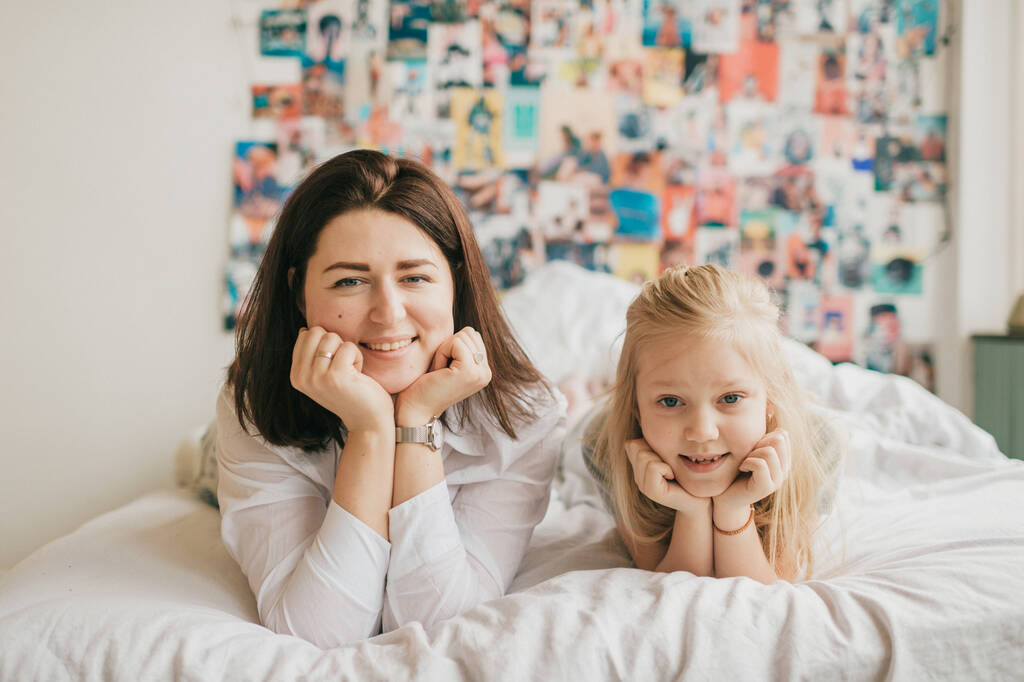 Styl życia Miękki portret ostrości szczęśliwej mamy przytula swoją uroczą córkę na białym łóżku. Wewnętrzny szczęśliwy rodzinny portret uśmiechniętej matki i jej córki leżących razem na łóżku z dekoracyjną ścianą za - Zdjęcie, obraz