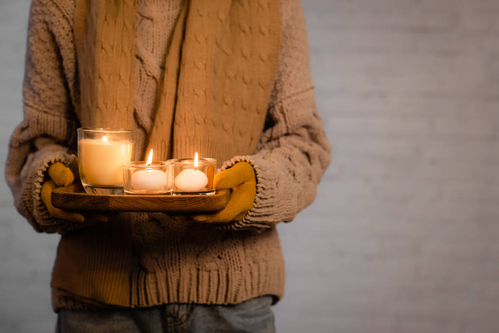 Vue recadrée des bougies brûlantes dans les mains de la femme en vêtements chauds et gants sur fond gris - Photo, image