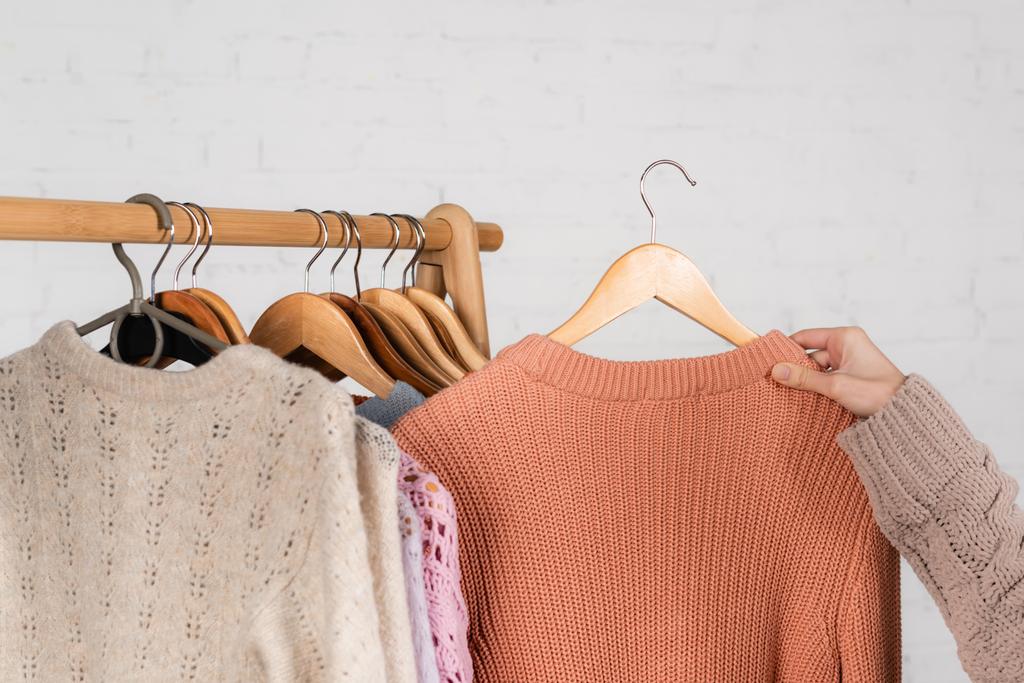 Cropped view of woman holding warm sweater near hanger rack on white background - Photo, Image