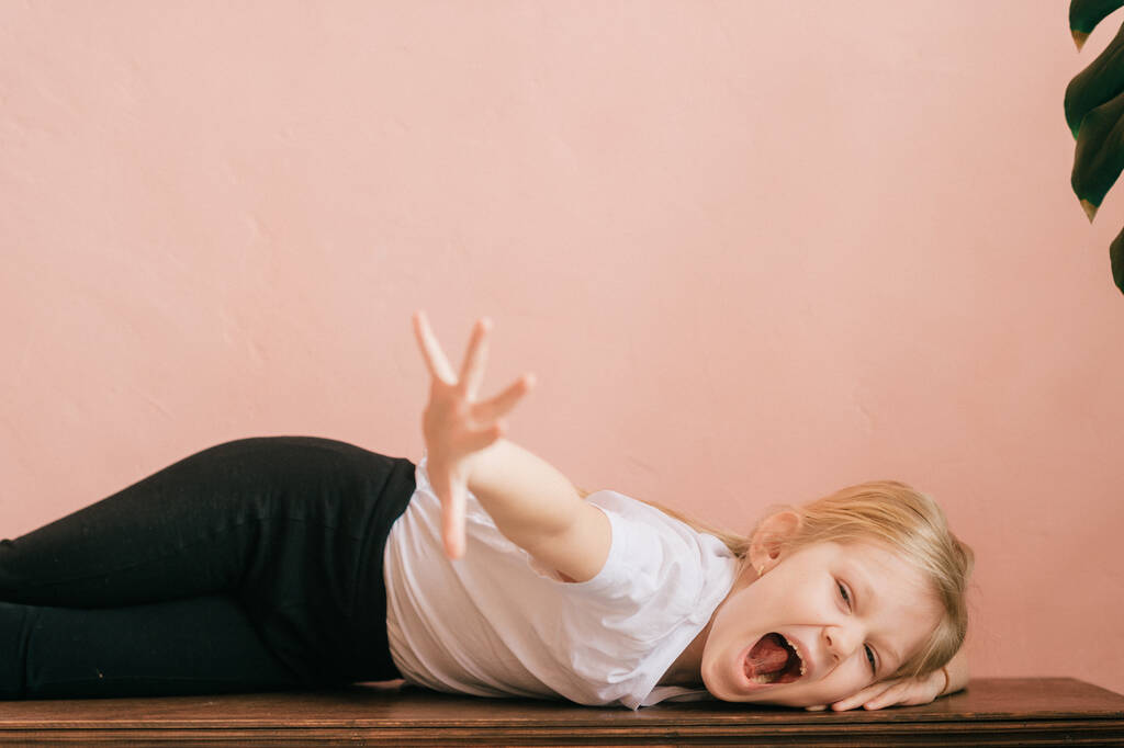Lifestyle portrait of clowning little girl make faces and shouting on pink wall background. Funny expressive female with ridiculous face kid lying on table and screaming like crazy with gesturing hand - Photo, Image