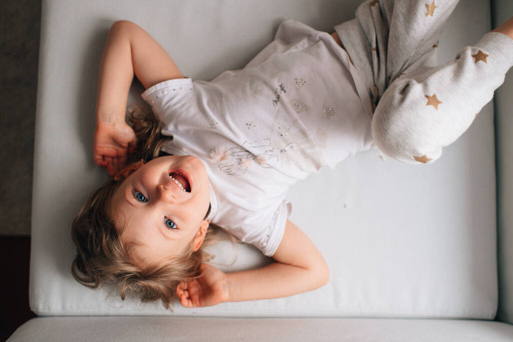 Πορτρέτο του χαριτωμένο μικρό καυκάσιο κορίτσι σε λευκό sleepwear βρίσκεται στο λευκό κρεβάτι της και χαίρεται - Φωτογραφία, εικόνα