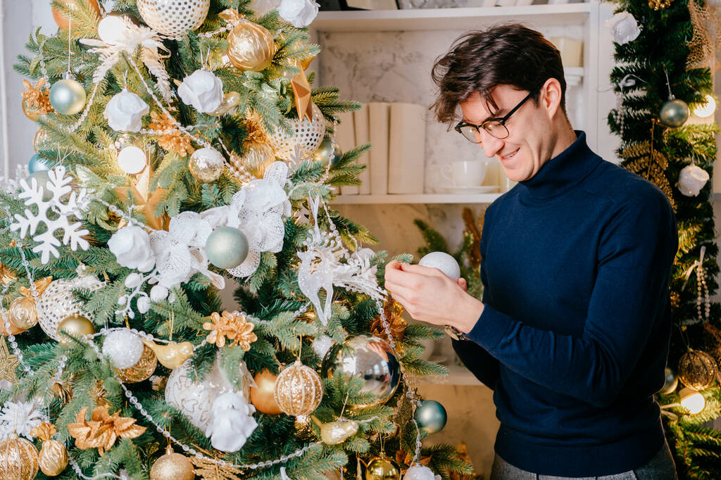 Feliz hombre guapo que adorna el árbol de Navidad en la habitación acogedora con decoraciones de vacaciones de año nuevo. - Foto, imagen