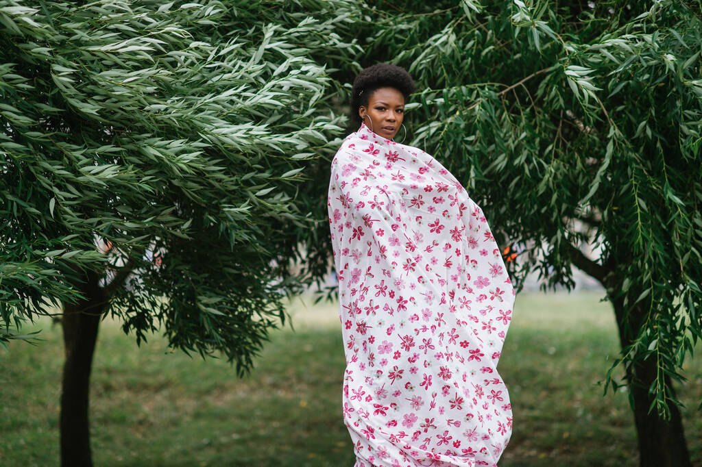 Gyönyörű sötét bőrű lány csomagolva lap divat nyomtatás pózol a parkban fák a háttérben. - Fotó, kép