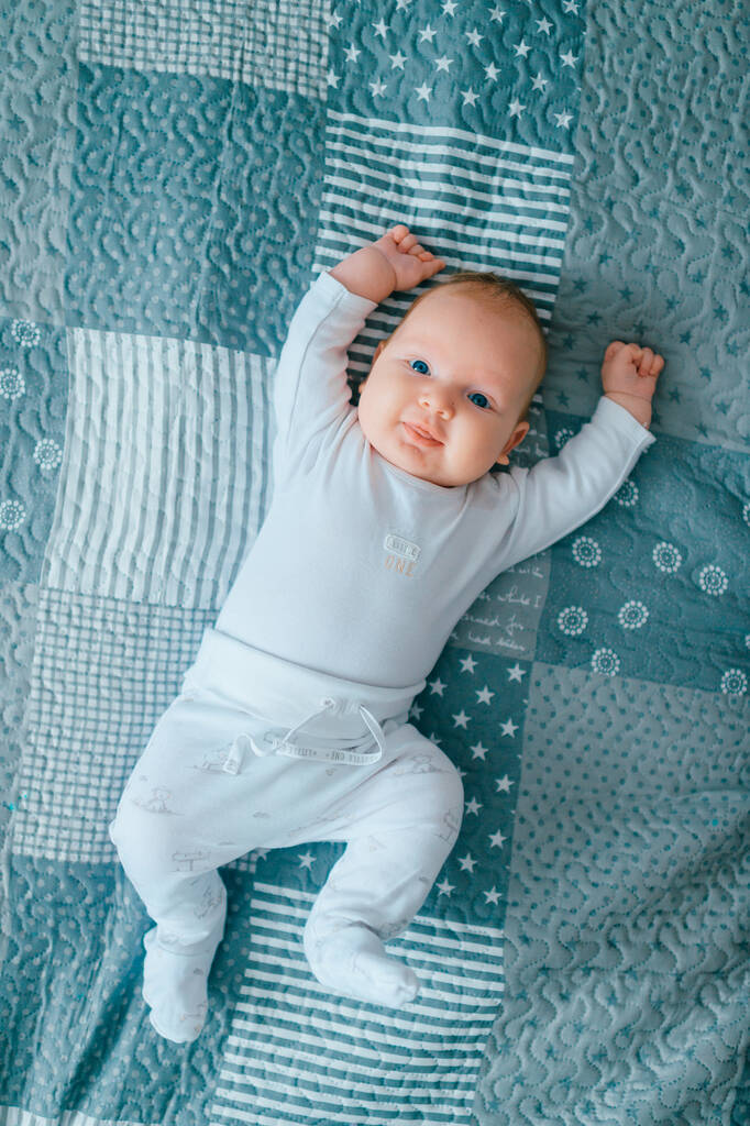 Imádnivaló újszülött férfi baba kék szemekkel életmód beltéri portré felülről. Aranyos gyerek, széttett kézzel, pijamában fekszik otthon, az ágyon. Kis gyermek érzelmi arc pihenés kanapén - Fotó, kép