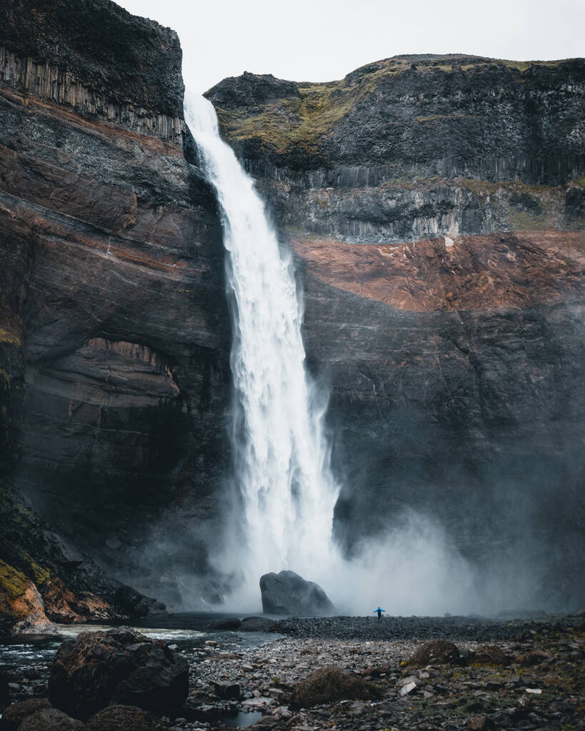 Een eenzame persoon die geniet van het prachtige uitzicht op de waterval omringd door rotsachtige kliffen in IJsland - Foto, afbeelding