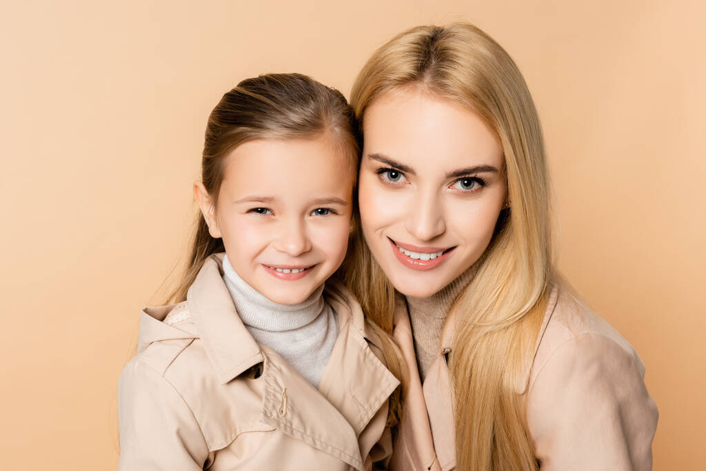 vrolijk blond moeder en gelukkig dochter in jassen kijken naar camera geïsoleerd op beige - Foto, afbeelding