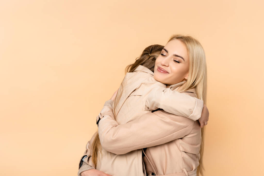 Ευτυχισμένη ξανθιά μητέρα αγκαλιάζει την κόρη της σε καμπαρντίνα απομονωμένη σε μπεζ - Φωτογραφία, εικόνα