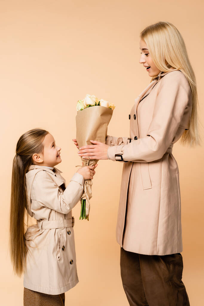 side view of cheerful kid giving flowers to surprised mother on 8 march isolated on beige - Photo, Image