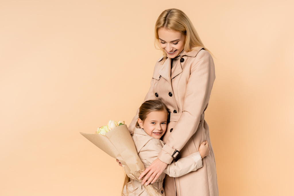 felice madre che tiene i fiori e abbraccia allegra figlia isolata sul beige - Foto, immagini