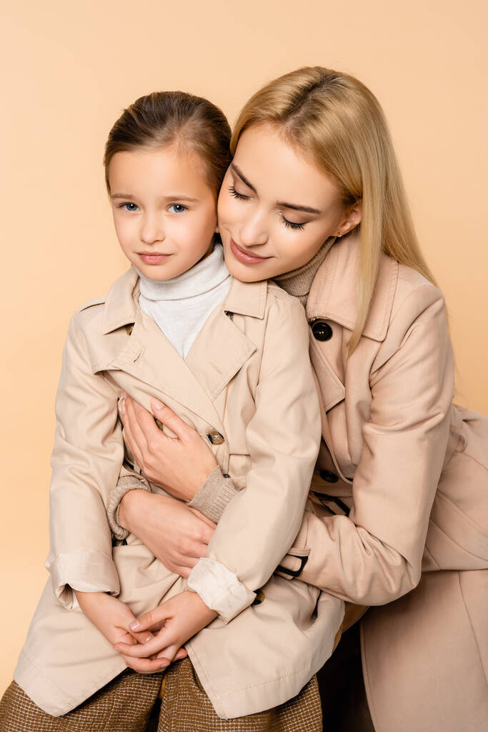 madre cariñosa abrazando hija en gabardina aislada en beige - Foto, imagen