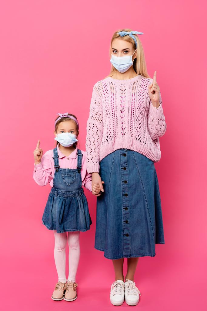 äidin ja tyttären täysi pituus lääketieteellisissä naamioissa, jotka osoittavat sormilla ja pitävät kädestä seisoessaan vaaleanpunaisella  - Valokuva, kuva