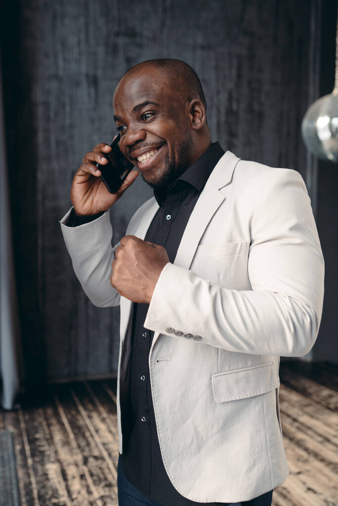 Açık ceketli ve siyah gömlekli Afro-Amerikan bir iş adamı telefonda konuşuyor ve anlaşmanın sonucuna seviniyor. - Fotoğraf, Görsel