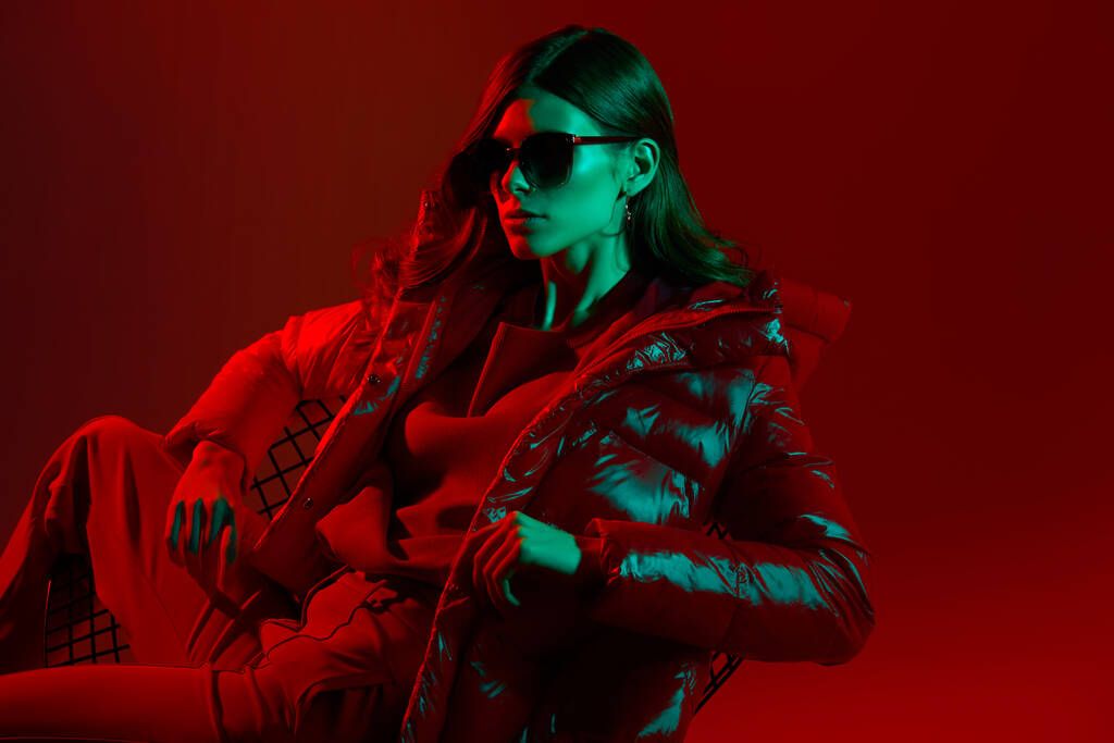 Fashion portrait of elegant trendy woman in red down jacket. Neon light, sunglasses, studio shot - Photo, Image
