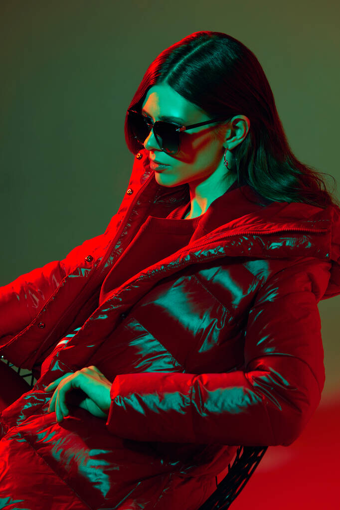Fashion portret van elegante trendy vrouw in rood donsjas. Neon licht, zonnebril, studio opname - Foto, afbeelding