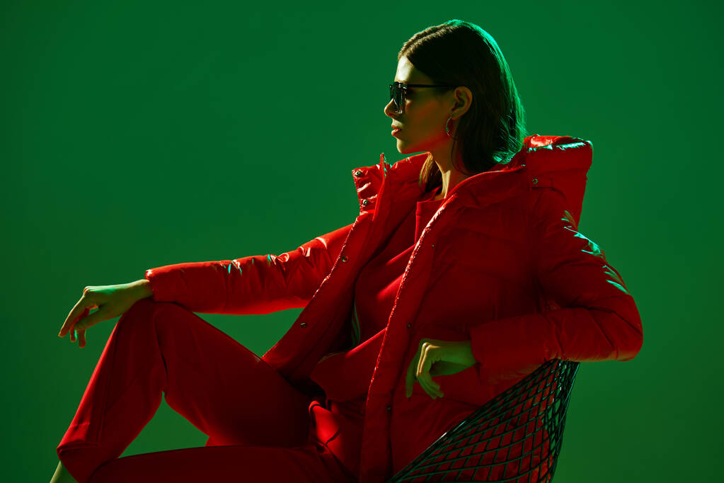 Modeporträt einer eleganten, trendigen Frau in roter Daunenjacke. Neonlicht, Sonnenbrille, Studioaufnahme - Foto, Bild