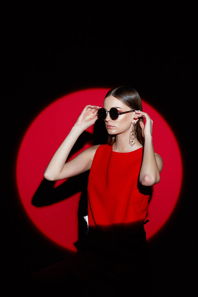 Kırmızı elbiseli genç bir kadının moda portresi. Güneş gözlüğü, kırmızı arkaplan, spot ışığı  - Fotoğraf, Görsel