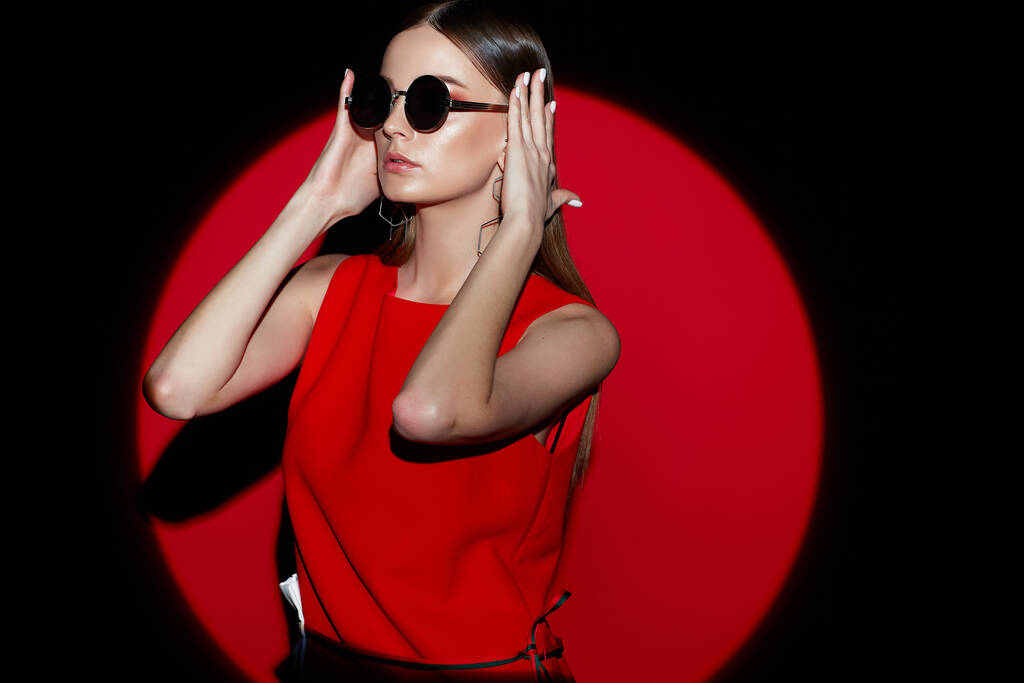 Kırmızı elbiseli genç bir kadının moda portresi. Güneş gözlüğü, kırmızı arkaplan, spot ışığı  - Fotoğraf, Görsel