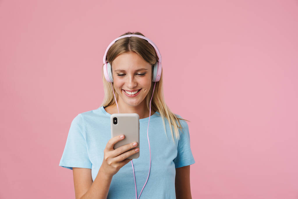 Chica rubia alegre en auriculares con teléfono móvil aislado sobre fondo rosa - Foto, imagen