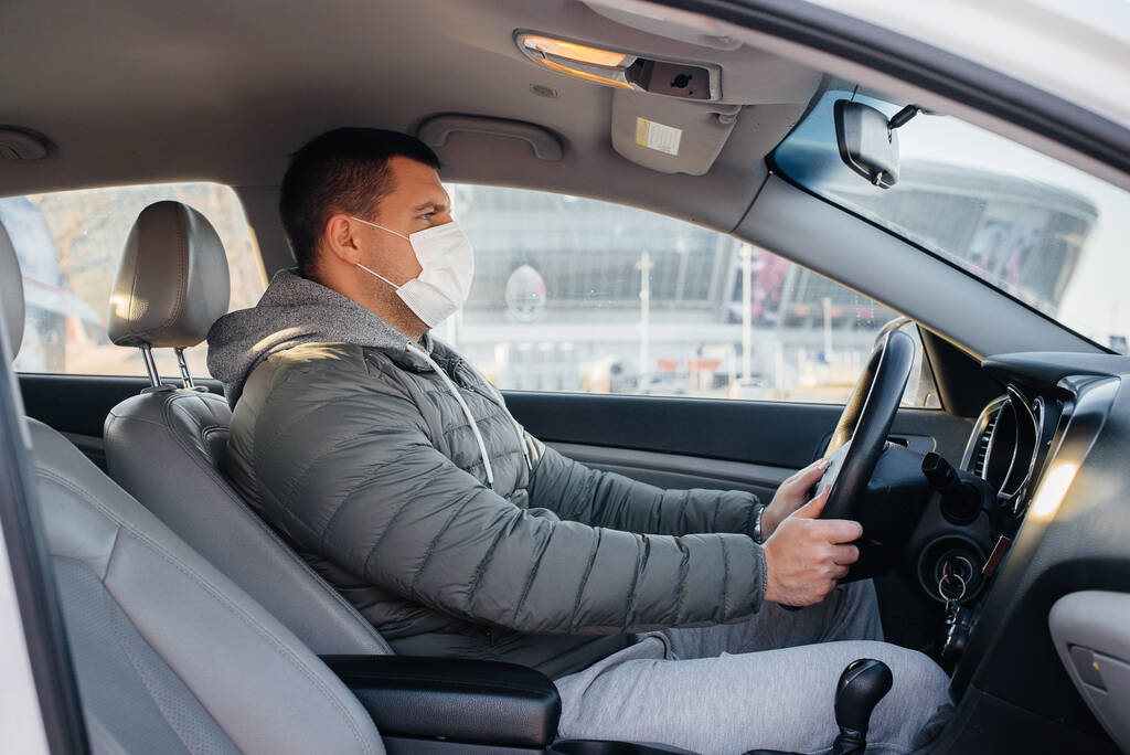 パンデミックとコロナウイルスの間、運転している間、若い男は個人的な安全のためにマスクをつけて車輪の後ろに座っています。流行 - 写真・画像
