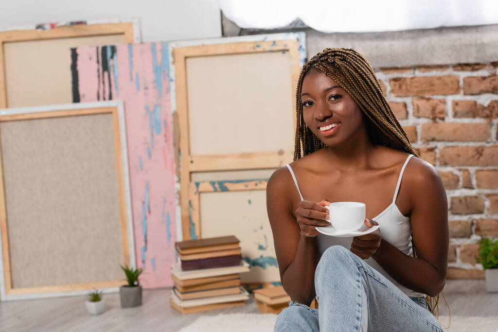 Χαμογελώντας αφροαμερικανή γυναίκα κρατώντας την κούπα του καφέ και κοιτάζοντας την κάμερα στο σπίτι  - Φωτογραφία, εικόνα