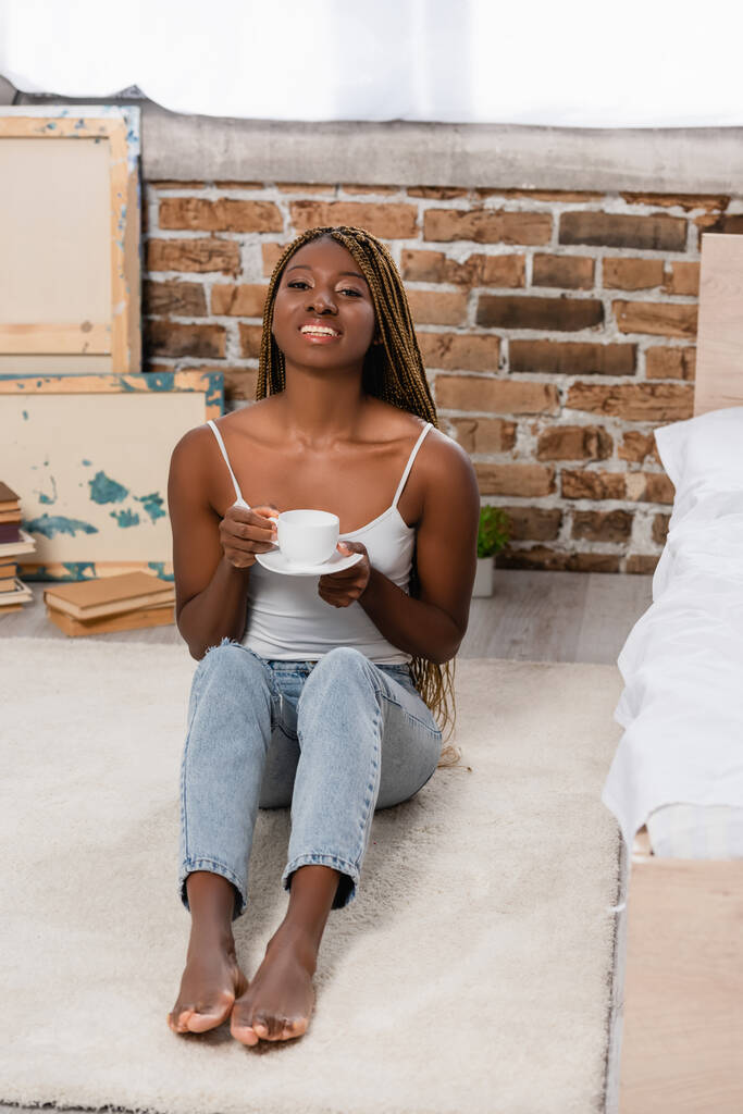 Vrolijke Afrikaanse Amerikaanse vrouw met kopje koffie terwijl ze op tapijt zit in de slaapkamer  - Foto, afbeelding
