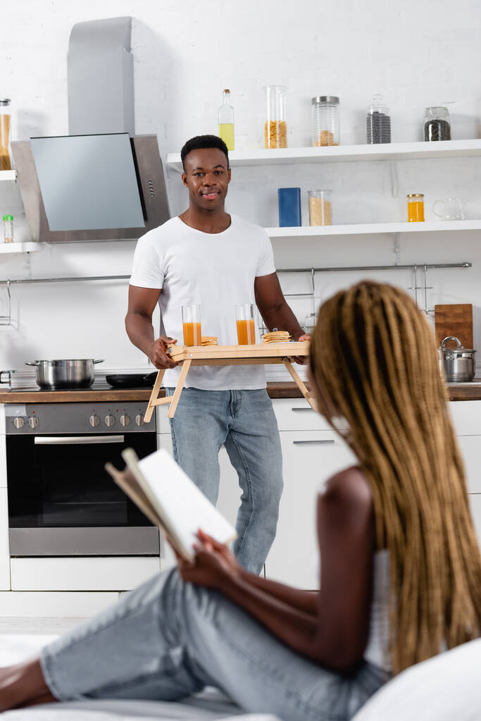 Smiling african man holding tray with tasty breakfast near girlfriend with book on bed on blurred foreground  - Photo, Image