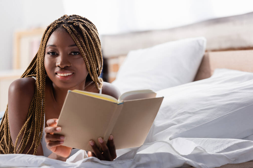Afričanky americká žena se usmívá na kameru, zatímco drží knihu na rozmazané popředí na posteli  - Fotografie, Obrázek