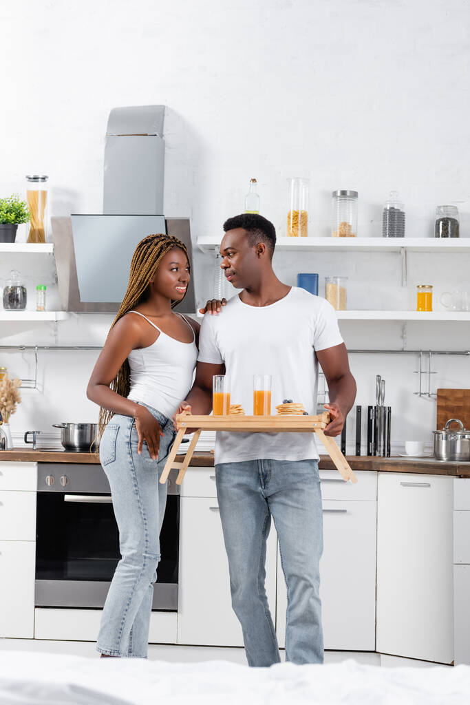 Smiling african american woman hugging boyfriend with orange juice and pancakes on tray  - Photo, Image