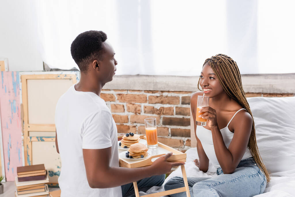 笑顔アフリカ系アメリカ人女性保持ガラスのオレンジジュース近くボーイフレンドとともにパンケーキオントレイでベッドルーム  - 写真・画像