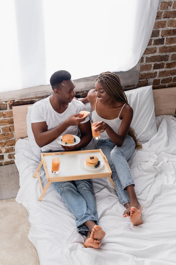 Высокий угол обзора улыбающейся африканской американки со стаканом апельсинового сока возле бойфренда с блином на кровати  - Фото, изображение
