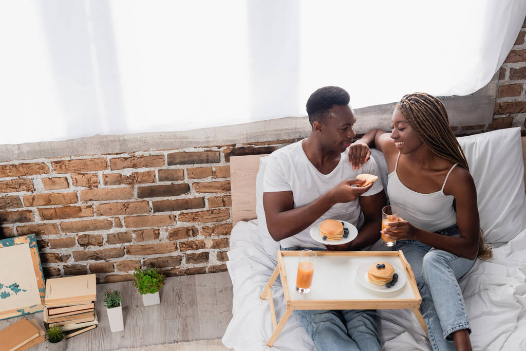 ベッドの上でおいしい朝食の近くのベッドに座っているアフリカ系アメリカ人のカップルの笑顔の高い角度ビュー  - 写真・画像