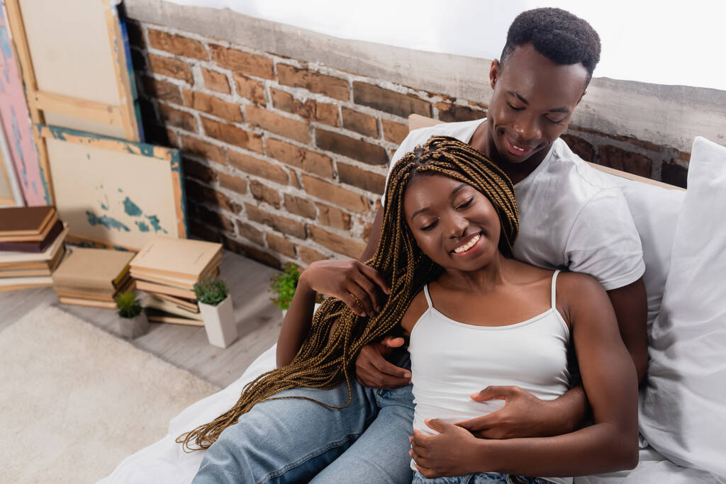 Vista de ángulo alto del hombre afroamericano sonriente abrazando a su novia con los ojos cerrados en la cama  - Foto, Imagen