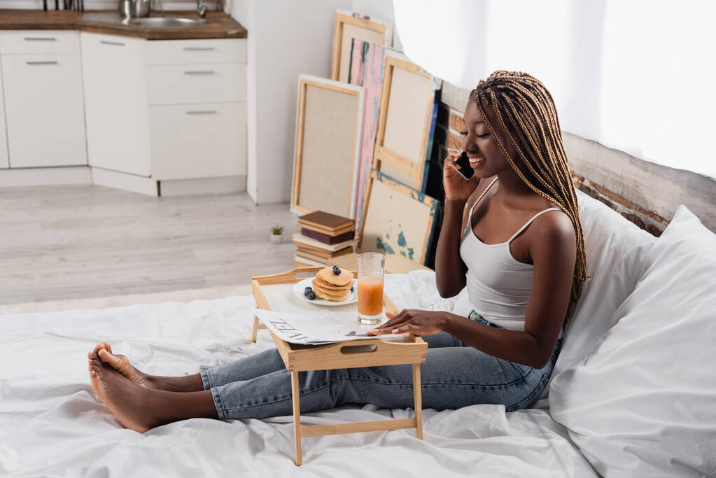 アフリカ系アメリカ人の女性が新聞の近くのスマートフォンで話しているのを笑って、ベッドの上でトレイで朝食  - 写真・画像
