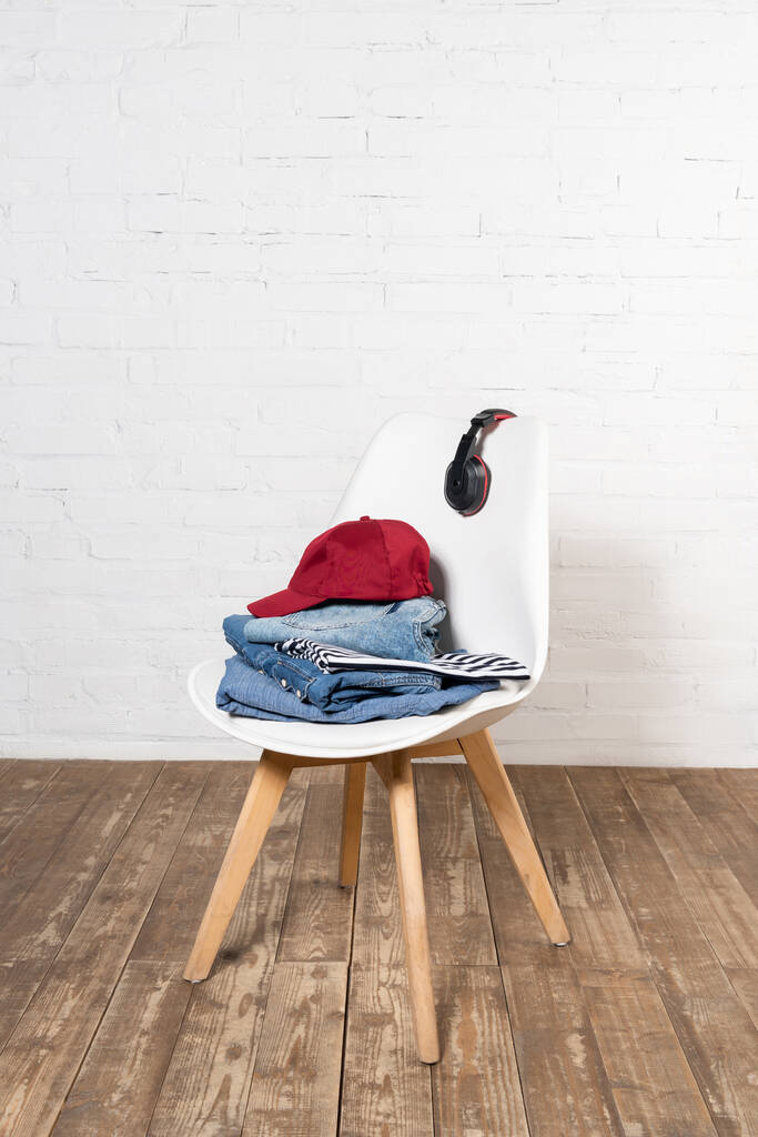 white chair with stacked denim clothes, burgundy cap and wireless headphones on wooden floor near brick wall - Photo, Image