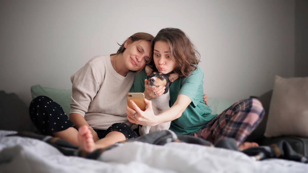 Όμορφη ευτυχισμένη λεσβιακό ζευγάρι κάθεται με πιτζάμες στο κρεβάτι το πρωί κάνοντας selfie στο smartphone με κατοικίδιο ζώο σκυλί bassengi - Φωτογραφία, εικόνα