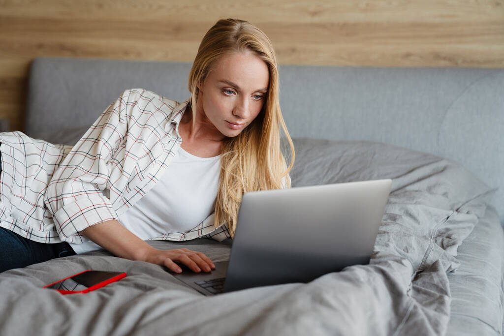 Schöne blonde fokussierte Frau benutzt Laptop, während sie zu Hause auf dem Bett liegt - Foto, Bild