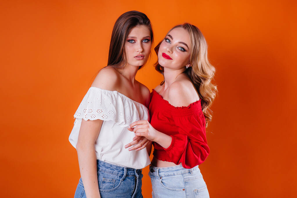 Zwei junge und schöne Mädchen zeigen Emotionen und lächeln im Studio auf orangefarbenem Hintergrund. Mädchen für die Werbung - Foto, Bild