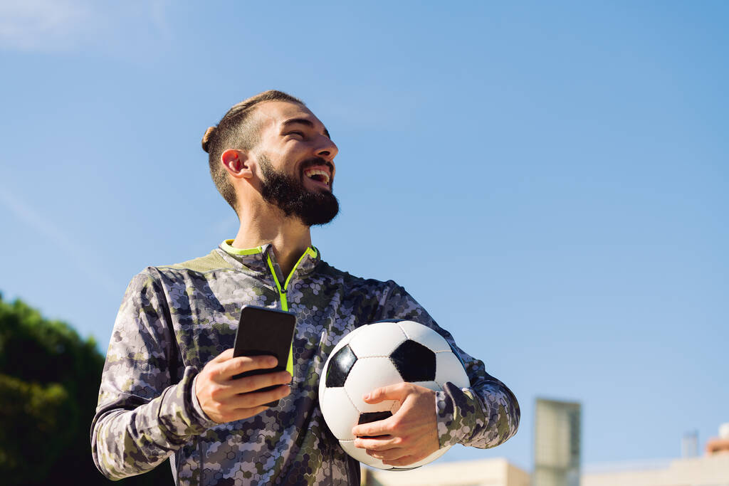 οριζόντια πορτρέτο του ένα χαρούμενο αθλητή με μια μπάλα ποδοσφαίρου γέλιο με το τηλέφωνο στο χέρι του, την έννοια της τεχνολογίας και του αστικού αθλητισμού τρόπο ζωής στην πόλη, αντίγραφο χώρο για κείμενο - Φωτογραφία, εικόνα