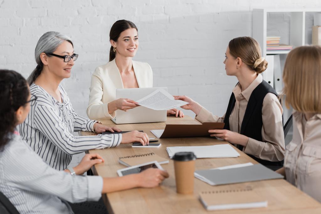 glücklicher Teamleiter übergibt Dokument an Mitarbeiterin in der Nähe multikultureller Geschäftsfrauen während eines Treffens - Foto, Bild