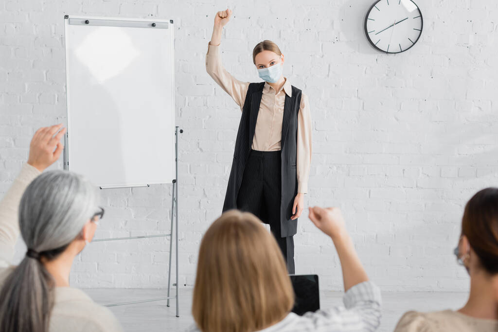 speaker in medical mask raising hand while standing near flipchart and looking at businesswomen on blurred foreground - Photo, Image